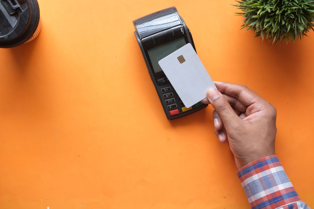 Vista cenital de una mano sosteniendo una tarjeta bancaria sobre un datafono en un escritorio naranja