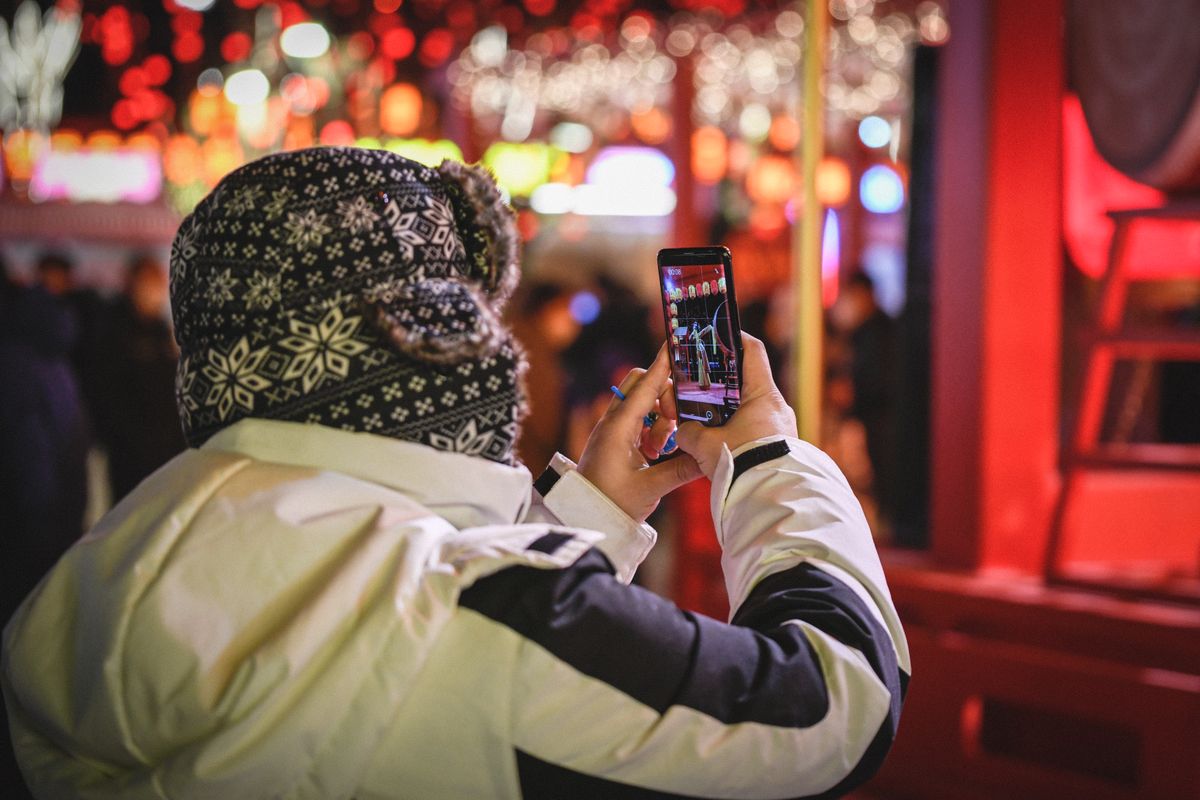 Viajero sosteniendo un smartphone para fotografiar una calle nocturna china iluminada con farolillos, la escena visible en la pantalla del teléfono