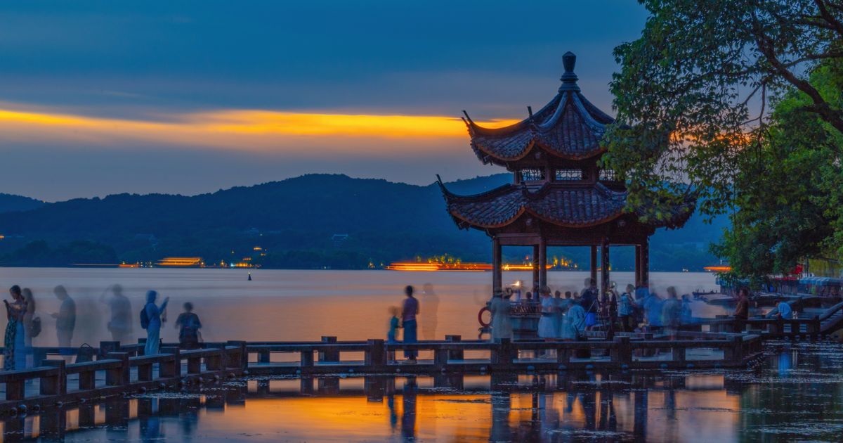 A pavilion on West Lake at sunset, with visitors gathered on the lakeside walkway