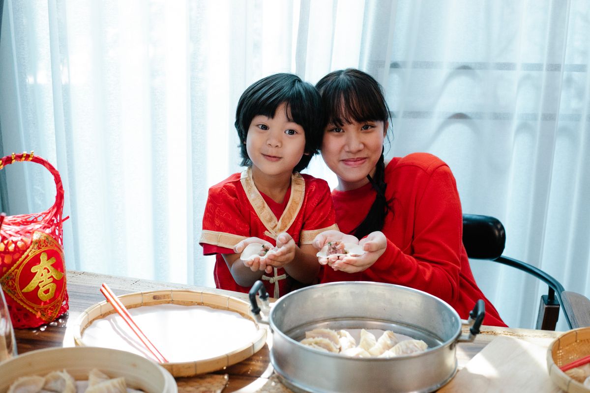 Two children in red clothing help fold dumplings on a table set with steamers for a Chinese New Year meal