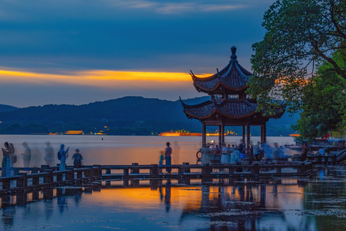 West Lake in Hangzhou at dusk with a lakeside pavilion and visitors along the shore