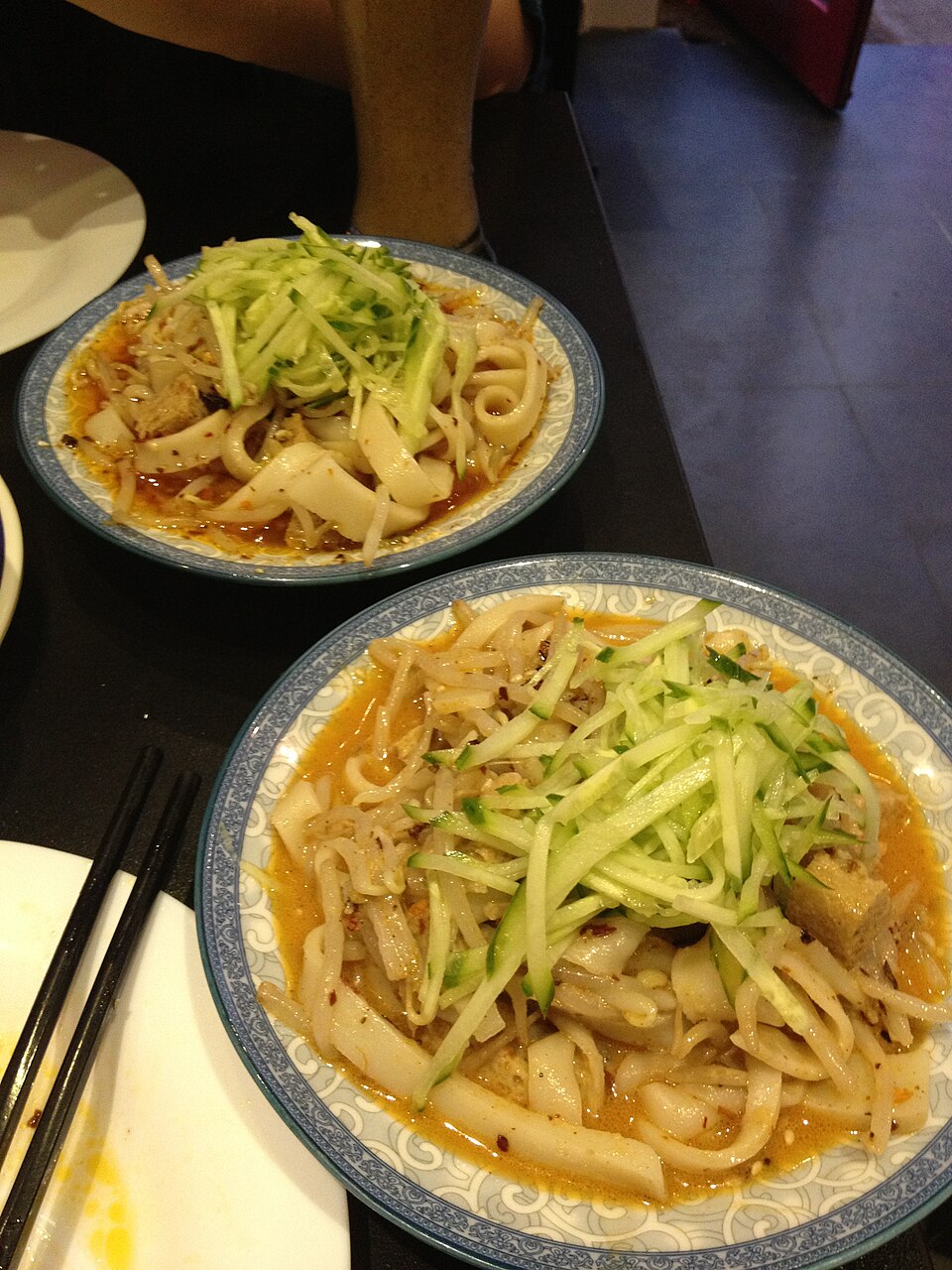 A bowl of cold noodles being topped with peanuts and cucumber